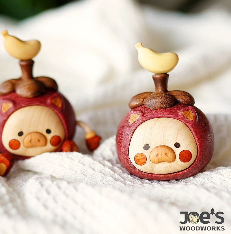 Two handmade wooden pig desk ornaments in fruit-themed designs resting on soft white fabric