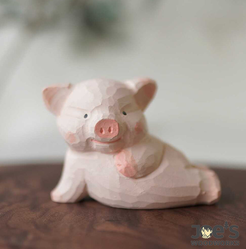 Hand-carved pink piglet wooden figurine smiling and laying on a table, handcrafted by Joe’s Woodworks.